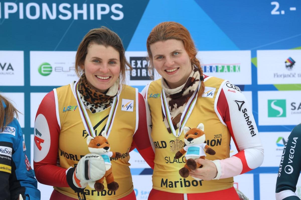 Two female athletes wearing a gold bib smile on the podium